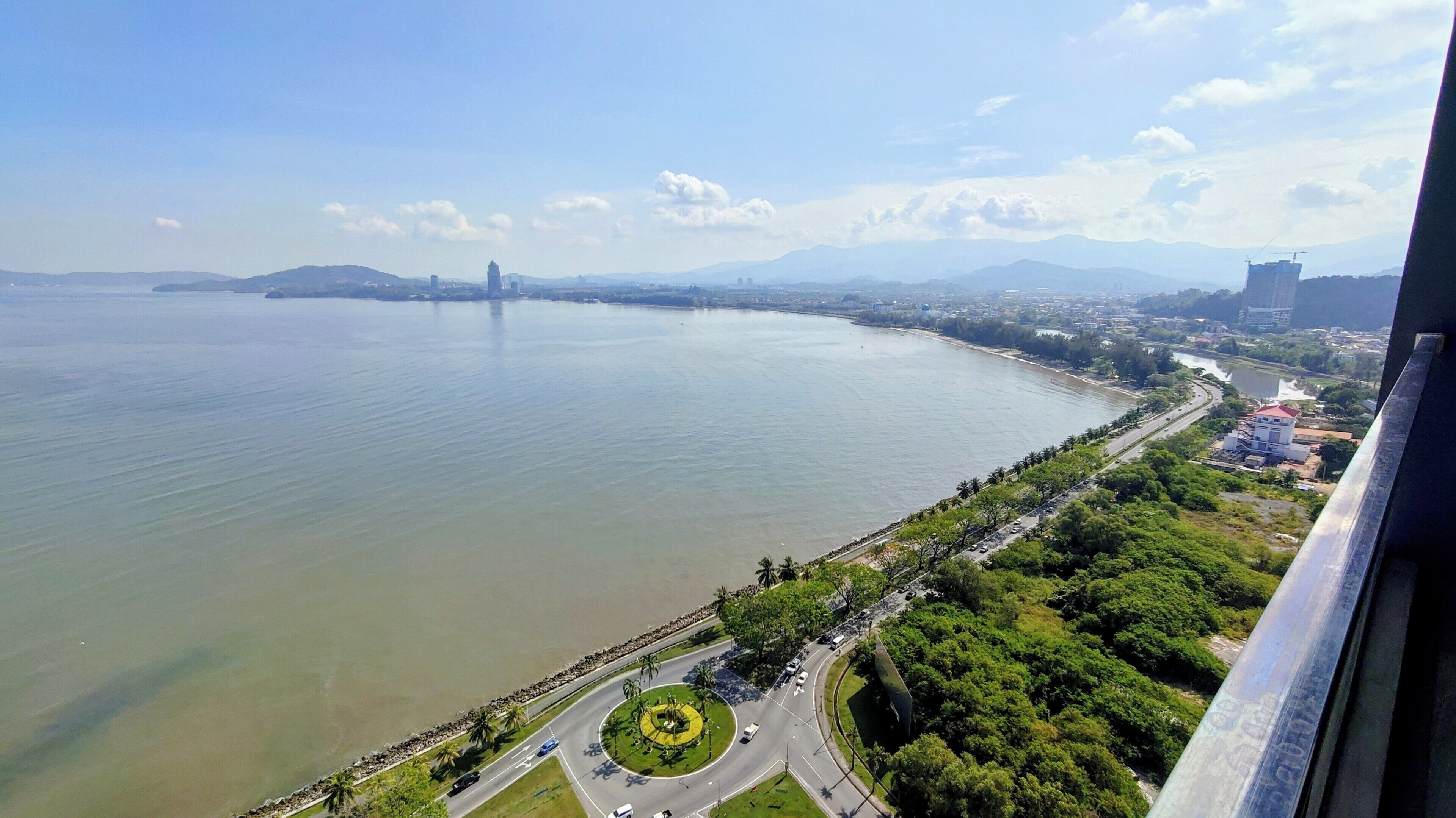 A panoramic view of a coastal city with a large body of water, a prominent bridge, and a well-maintained park with a circular fountain.