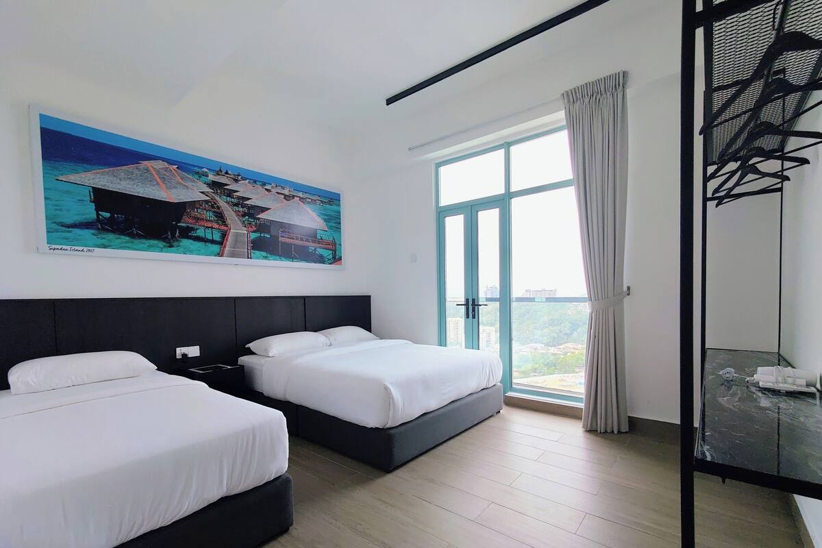 A hotel room with two beds, a large window with a view of a cityscape, a painting of a tropical scene, and a black metal structure on the right.