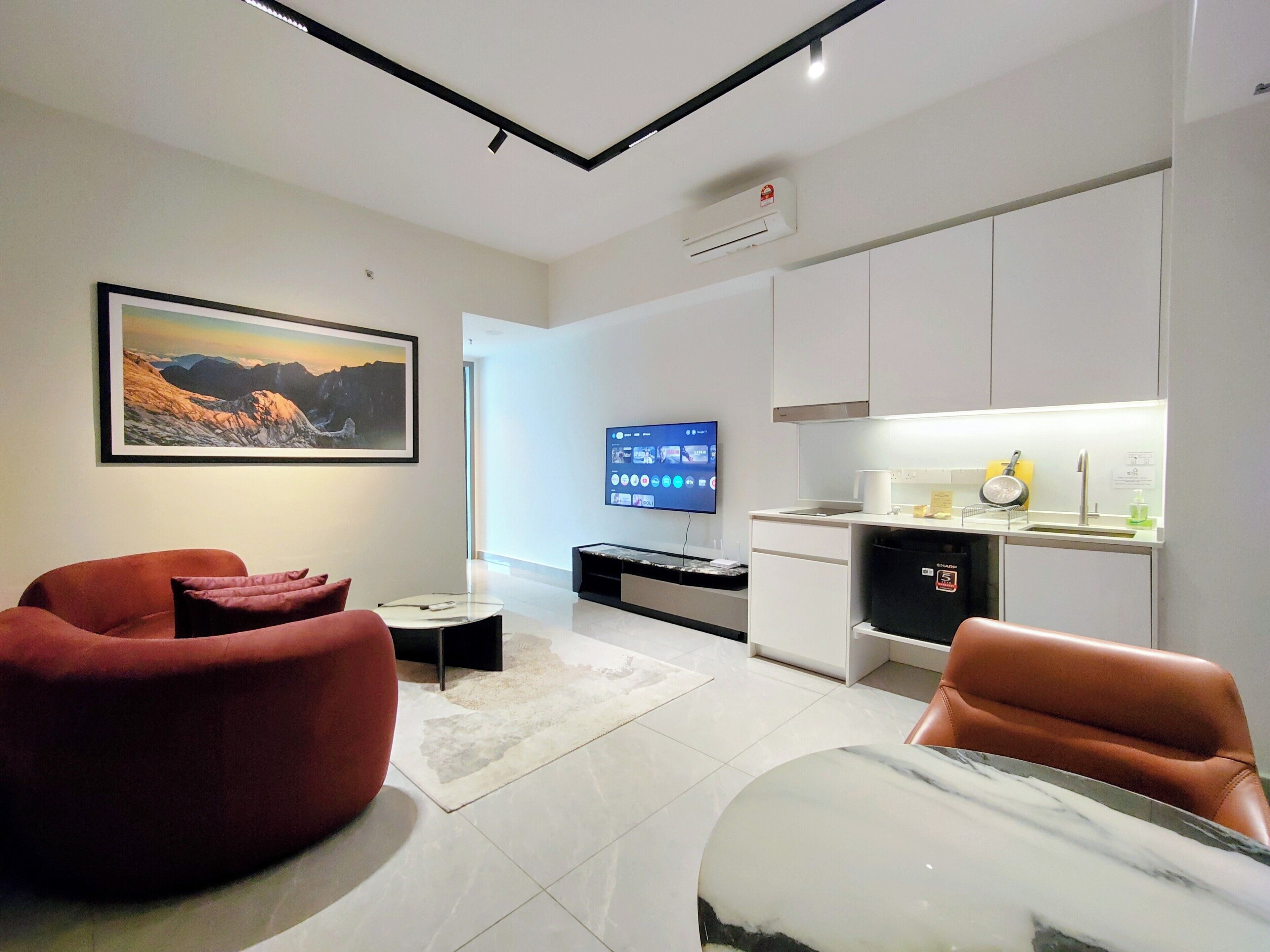 Een moderne woonkamer met een flatscreen televisie, rode banken, een marmeren salontafel en een keuken met een gootsteen en fornuis.
