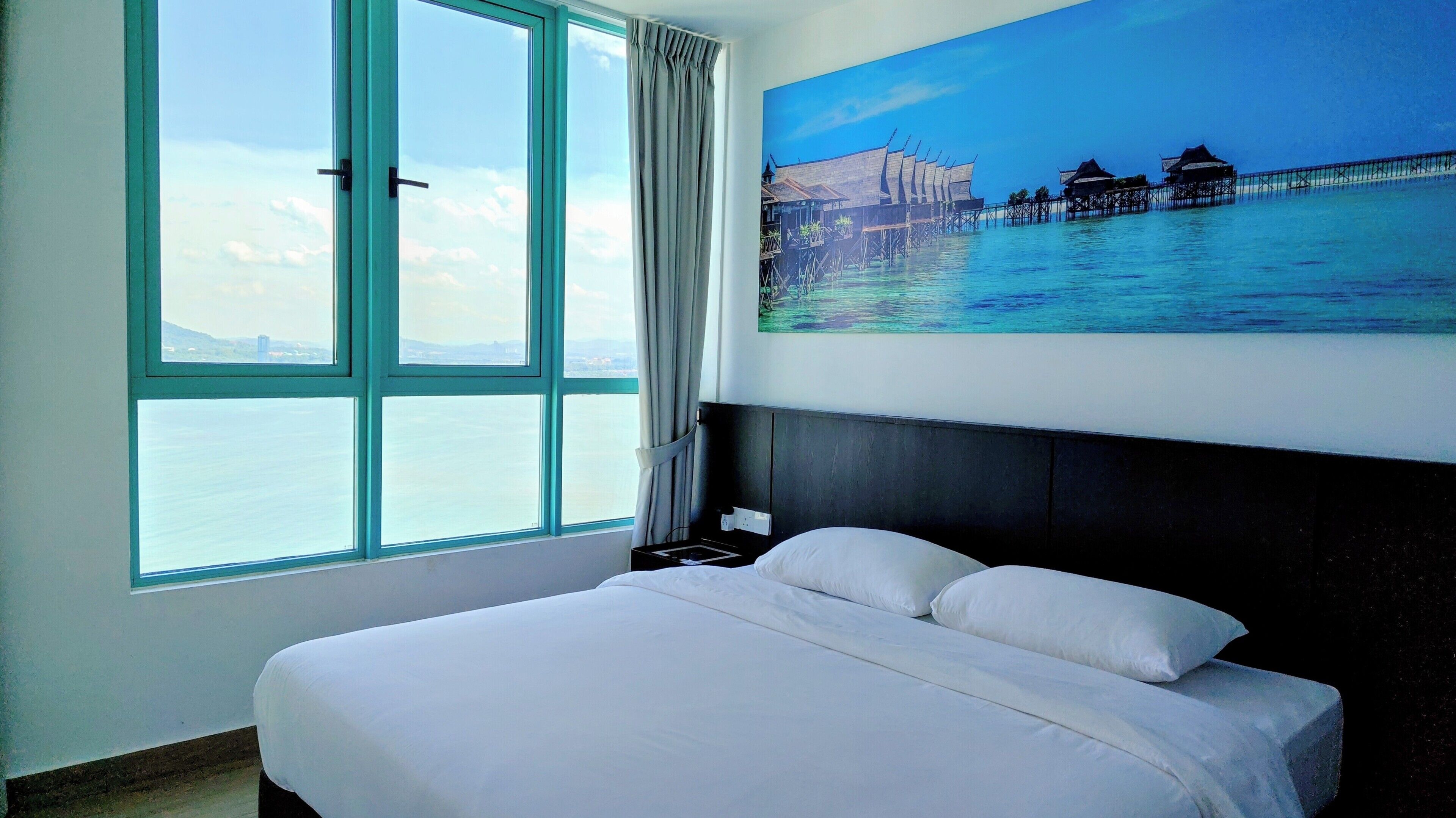 A hotel room with a large bed, a painting of a coastal scene, and a view of the sea through a window.