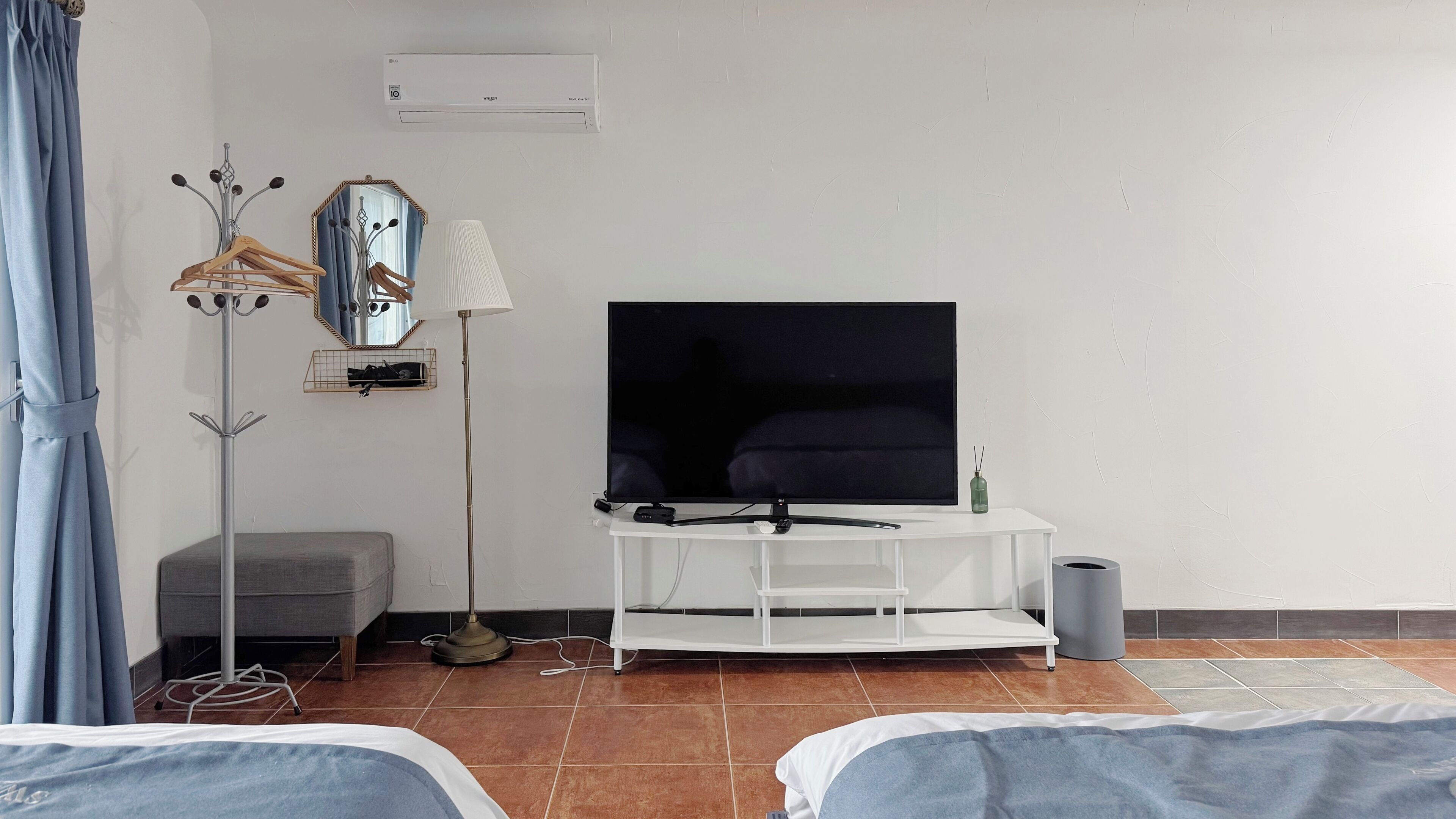 A hotel room with a TV on a white stand, a grey sofa, a small table with a lamp, and a mirror on the wall.