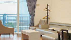 A modern living room with a sofa, armchair, and a small table. A piano is mounted on the wall, and a balcony with a railing is visible through the window. - Sweet Stay (Seogwipo)