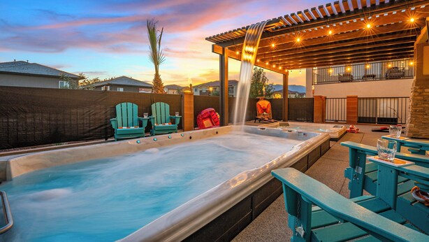 A hot tub with two Adirondack chairs, a pergola with string lights, and a view of a residential area. - Red Rock Roamin' | Private Pool, & Hot Tub | Resort Amenities | Sleeps 21 (Hurricane)