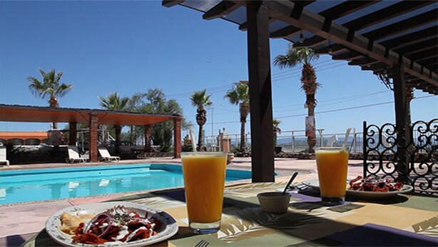 A poolside dining area with a table set for two, a pool, and palm trees in the background. - Hotel Villa Vitta (Bahia de los Angeles)