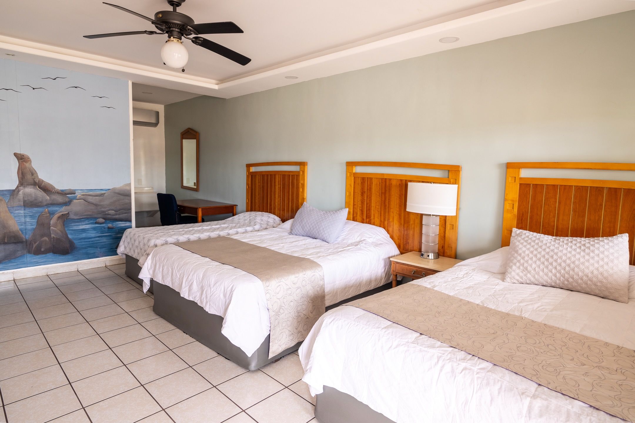 Habitación de hotel con tres camas, ventilador de techo, escritorio y un cuadro grande de lobos marinos.