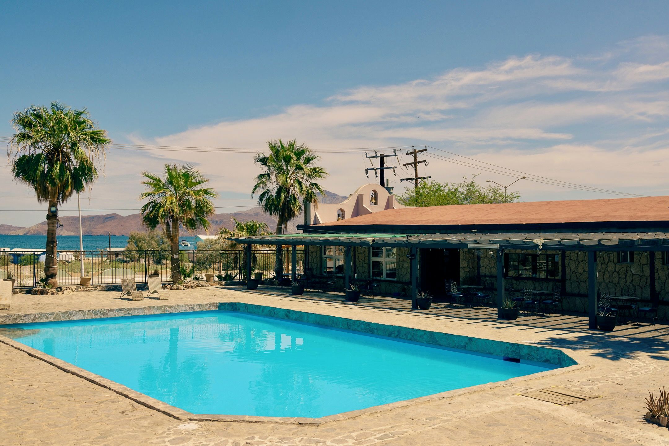 Una piscina con plataforma de piedra, palmeras y un edificio con techo rojo.