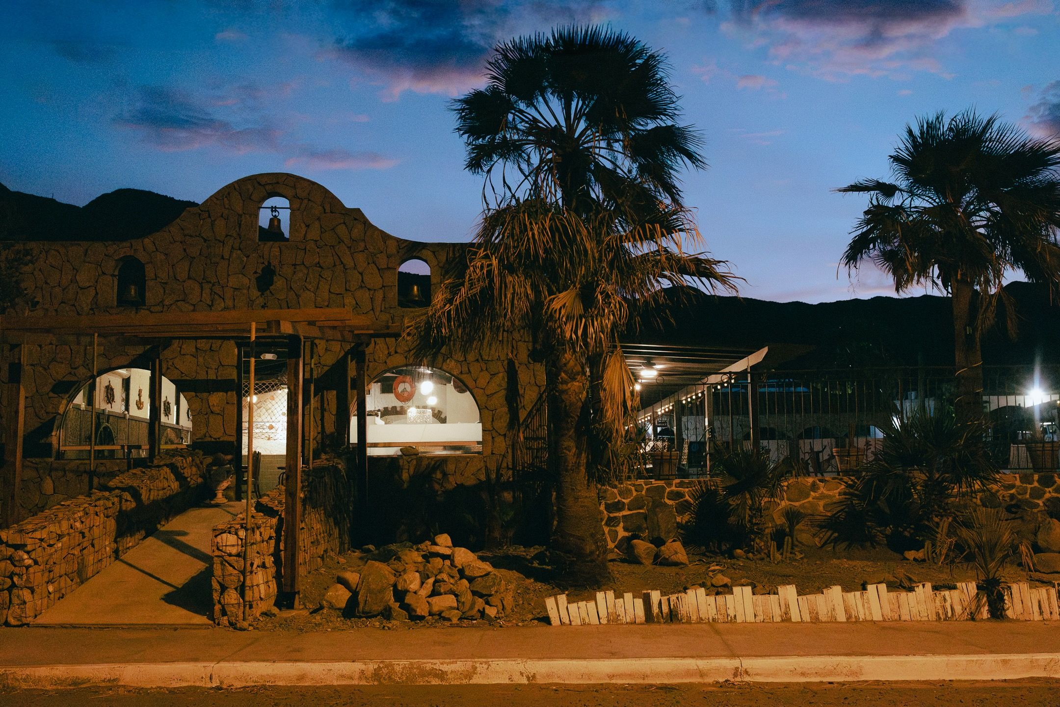 A stone building with a covered entrance, palm trees, and a mountainous backdrop.