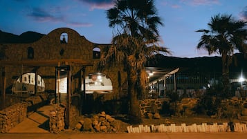 A stone building with a covered entrance, palm trees, and a mountainous backdrop.