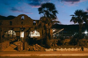 Un edificio de piedra con un porche cubierto, palmeras y montañas al fondo.