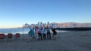 A group of people posing in front of a sign on a beach with chairs and a view of mountains in the background. - Hotel Villa Vitta (Bahia de los Angeles)