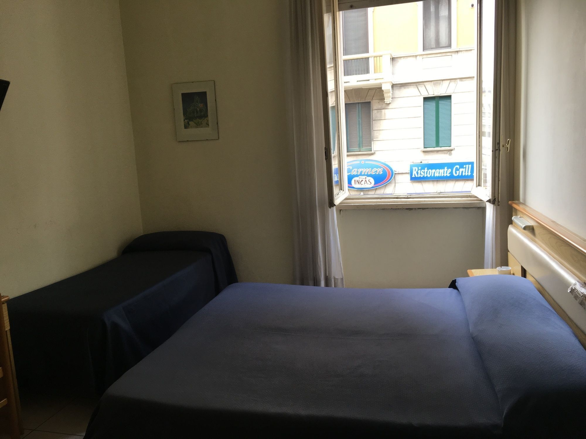 A hotel room with two beds, a window with a view of buildings, and a sign for Carmen Ristorante Grill.