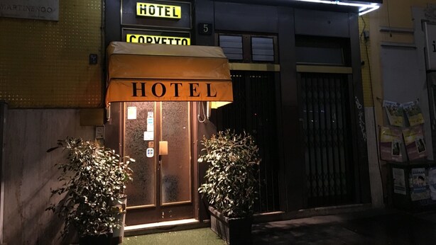 Exterior del Hotel Corvetto con un letrero de neón, una puerta de entrada visible y dos plantas en macetas al frente.