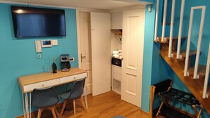 Una habitación pequeña con escritorio, dos sillas, cafetera, botella de vino, televisor, teléfono y una escalera.