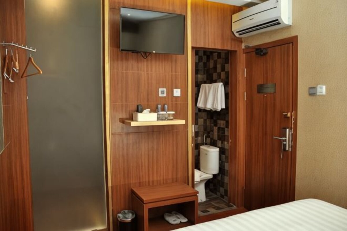 Ein Hotelzimmer mit einem Holzschrank, einem an der Wand befestigten Fernseher, einem kleinen Tisch mit Toilettenartikeln, einer Toilette und einem Bett.