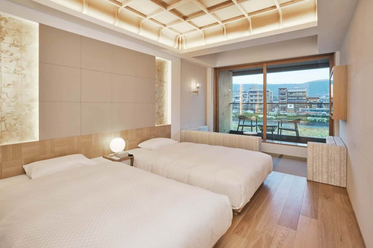 A modern hotel room with two beds, a large window with a city view, and a ceiling with a geometric light fixture.
