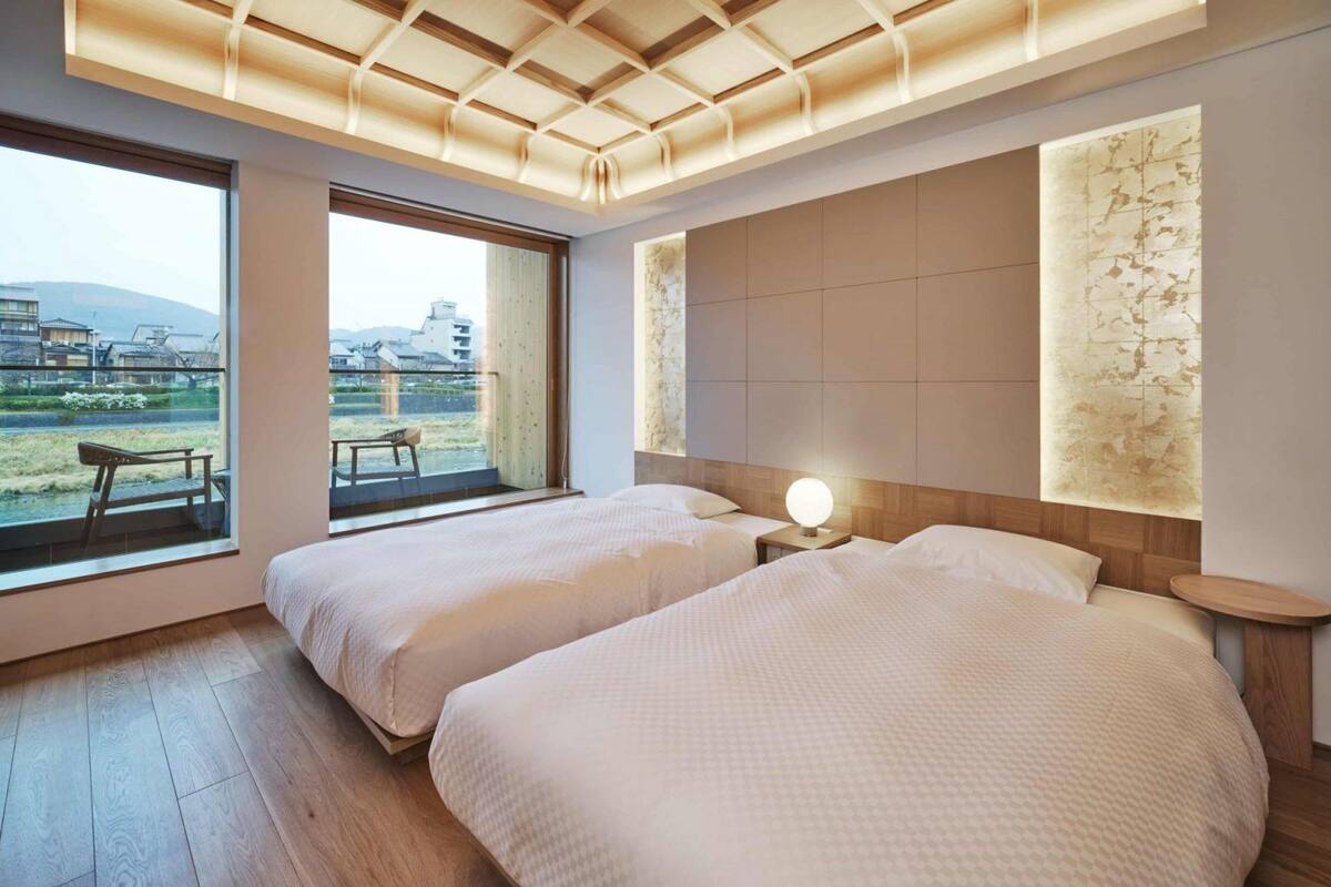 A hotel room with two beds, a large window with a view of the outdoors, a wooden ceiling, and a wall-mounted light fixture.