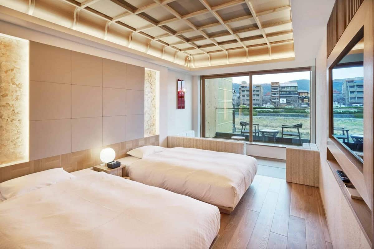 A hotel room with two beds, a large window with a view of buildings, and a ceiling with a wooden pattern.