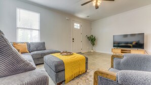 Un salon avec un canapé gris, un pouf et un fauteuil, un plaid jaune, un téléviseur posé sur un meuble en bois et un ventilateur de plafond.