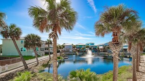 A pool with a fountain, surrounded by palm trees and houses with light blue and white exteriors.