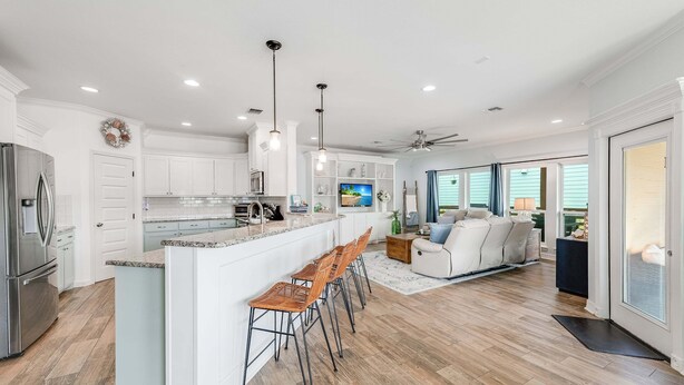A modern kitchen with white cabinetry, a central island with bar stools, a dining area with a sofa and chairs, and a large window providing natural light.