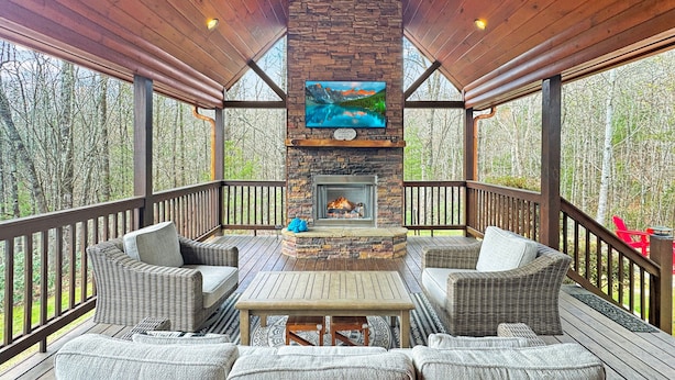 A covered outdoor deck with a stone fireplace, wicker furniture, and a view of the forest. - Mountain Solace - Pet Friendly | Hot Tub | Shuffleboard and Arcade Games (Blue Ridge)