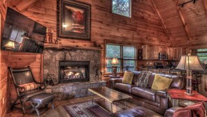 A cozy log cabin interior with a stone fireplace, leather furniture, and a kitchen area.
