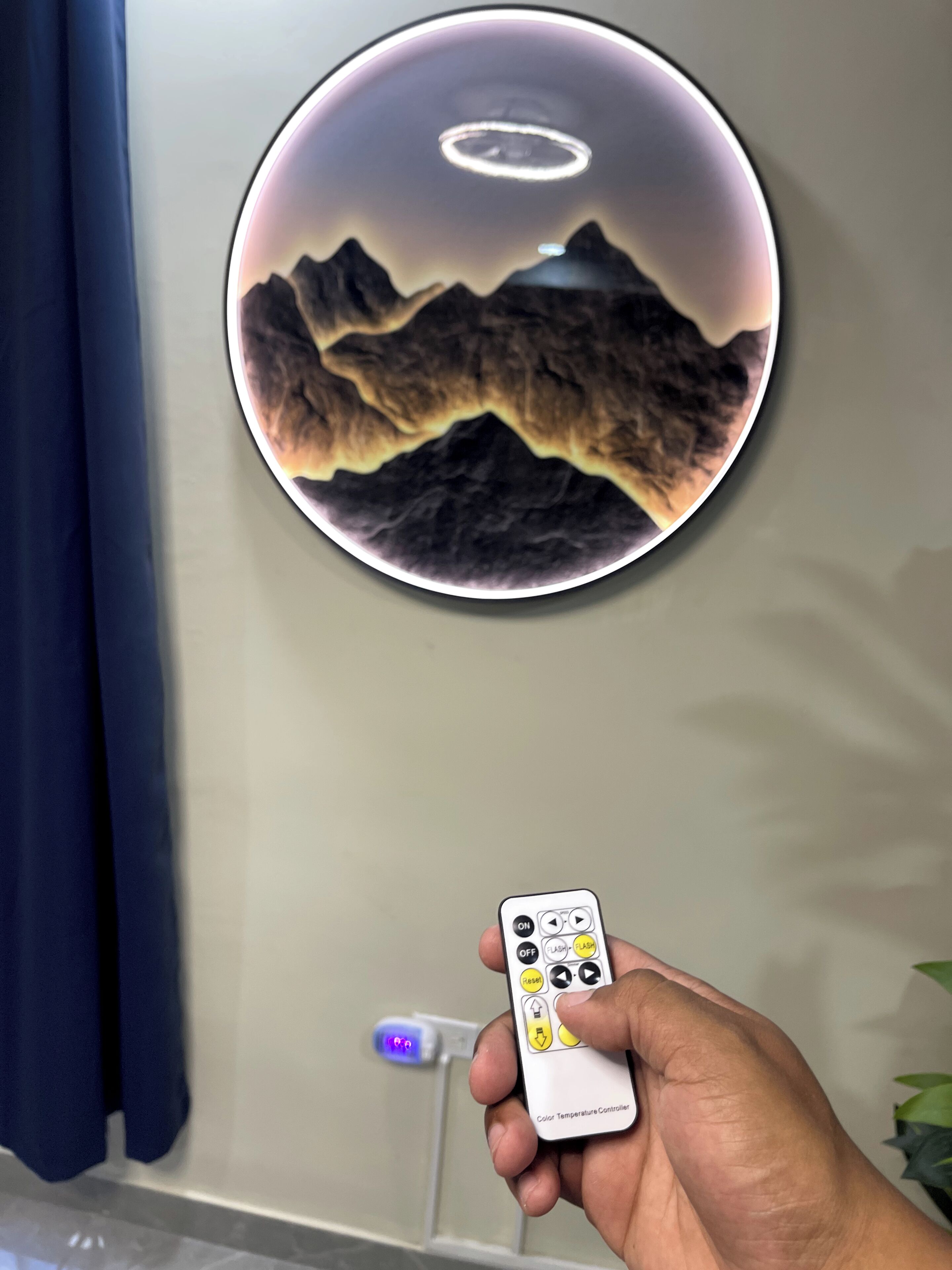 Een hand houdt een afstandsbediening vast, met een ronde wanddecoratie van bergen en een halo.