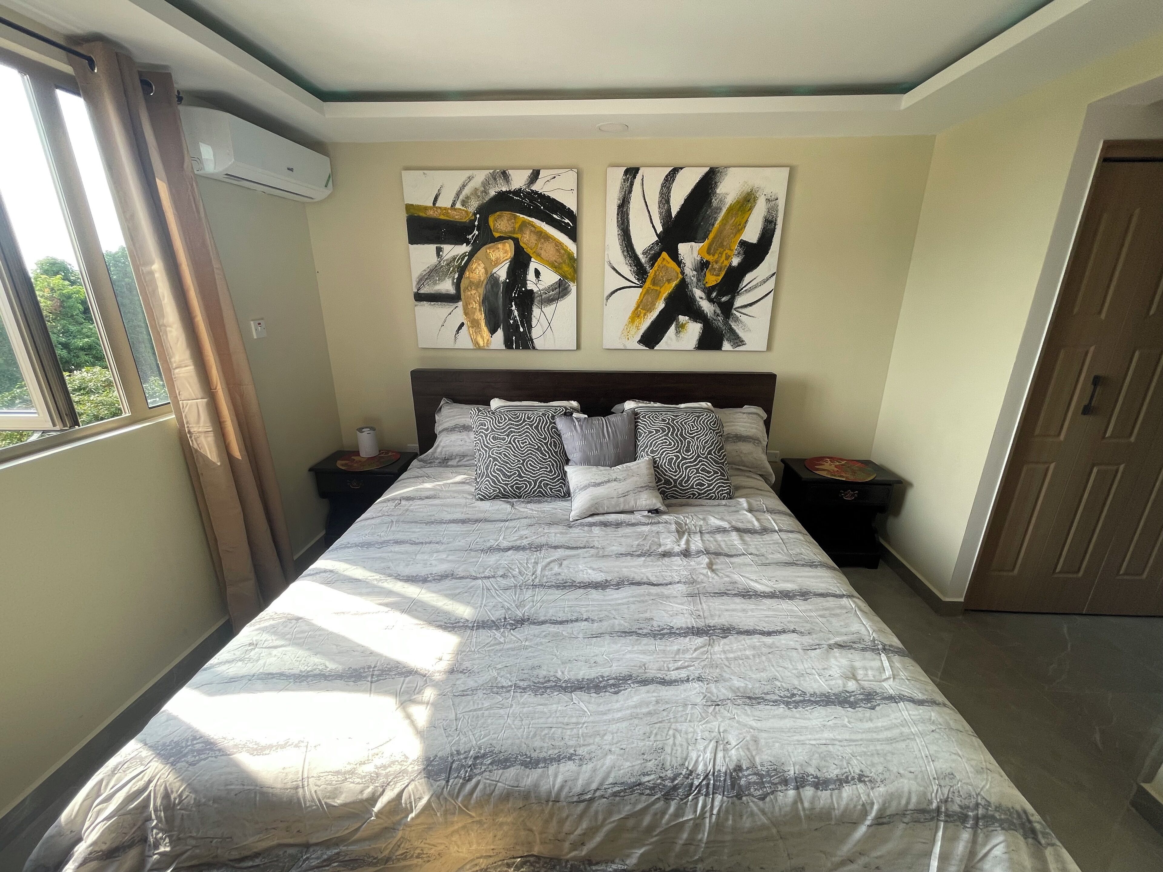 Ein Schlafzimmer mit einem großen Bett, zwei Nachttischen, einem Fenster mit Vorhängen und zwei abstrakten Bildern an der Wand.