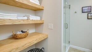 A bathroom with wooden shelves holding towels, a wicker basket, and a glass shower door. - Two-Home Heart of Austin Compound and Karaoke Room (Austin)