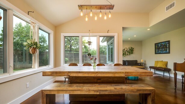 A spacious dining area with a large wooden table and bench, large windows, and a hanging light fixture. - Two-Home Heart of Austin Compound and Karaoke Room (Austin)