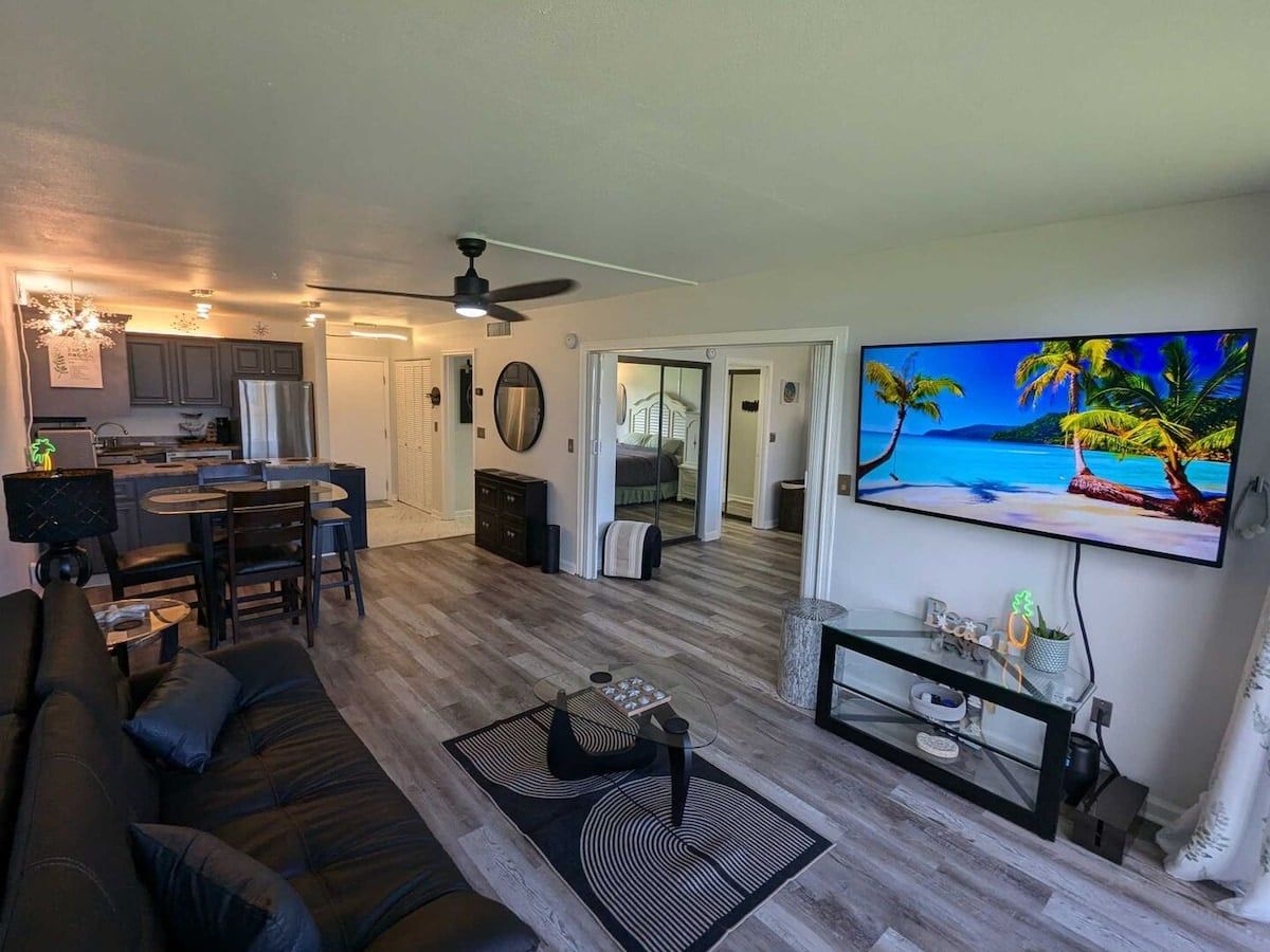 A modern living room with a sofa, coffee table, and TV. There is a kitchen area with cabinets and a dining table. The room has a ceiling fan and a large window.