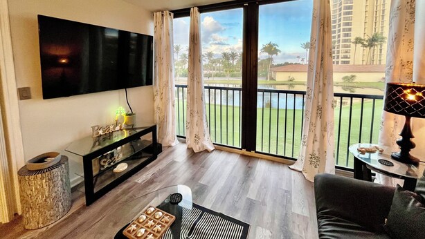 En stue med et fladskærms-tv, et sort konsolbord, en vævet kurv, en bakke med småkager, et sort sofabord, en sofa og udsigt til en balkon med pool og palmer.