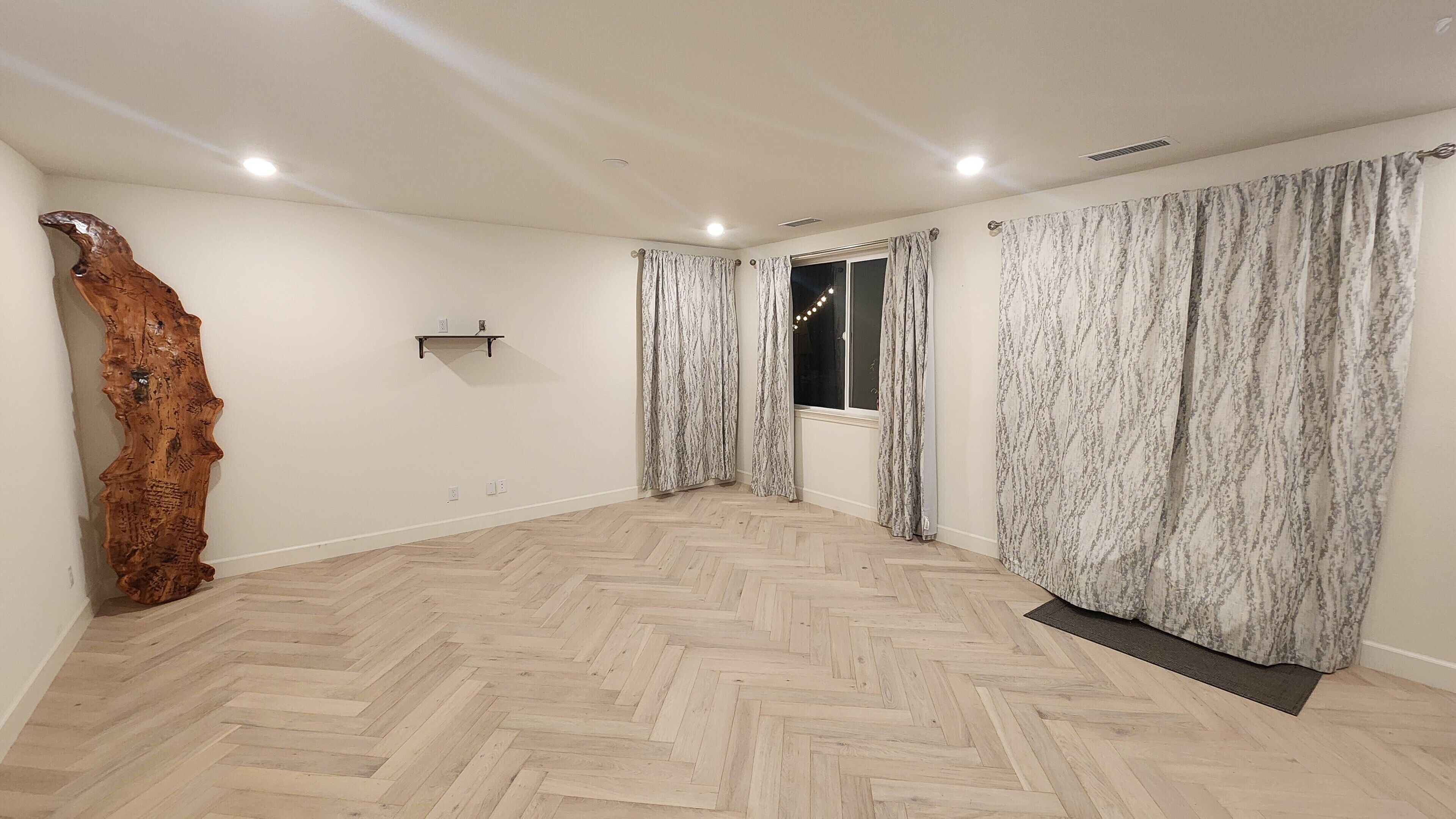Une pièce spacieuse dotée d’un parquet en bois à motifs chevrons, d’une grande sculpture en bois et d’une fenêtre avec des rideaux.
