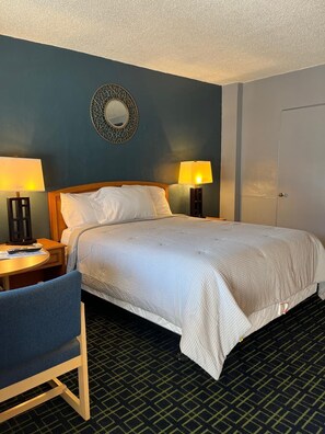 A hotel room with a bed, two bedside lamps, a round mirror, a desk with a chair, and a door. - American Inn (Sacramento)