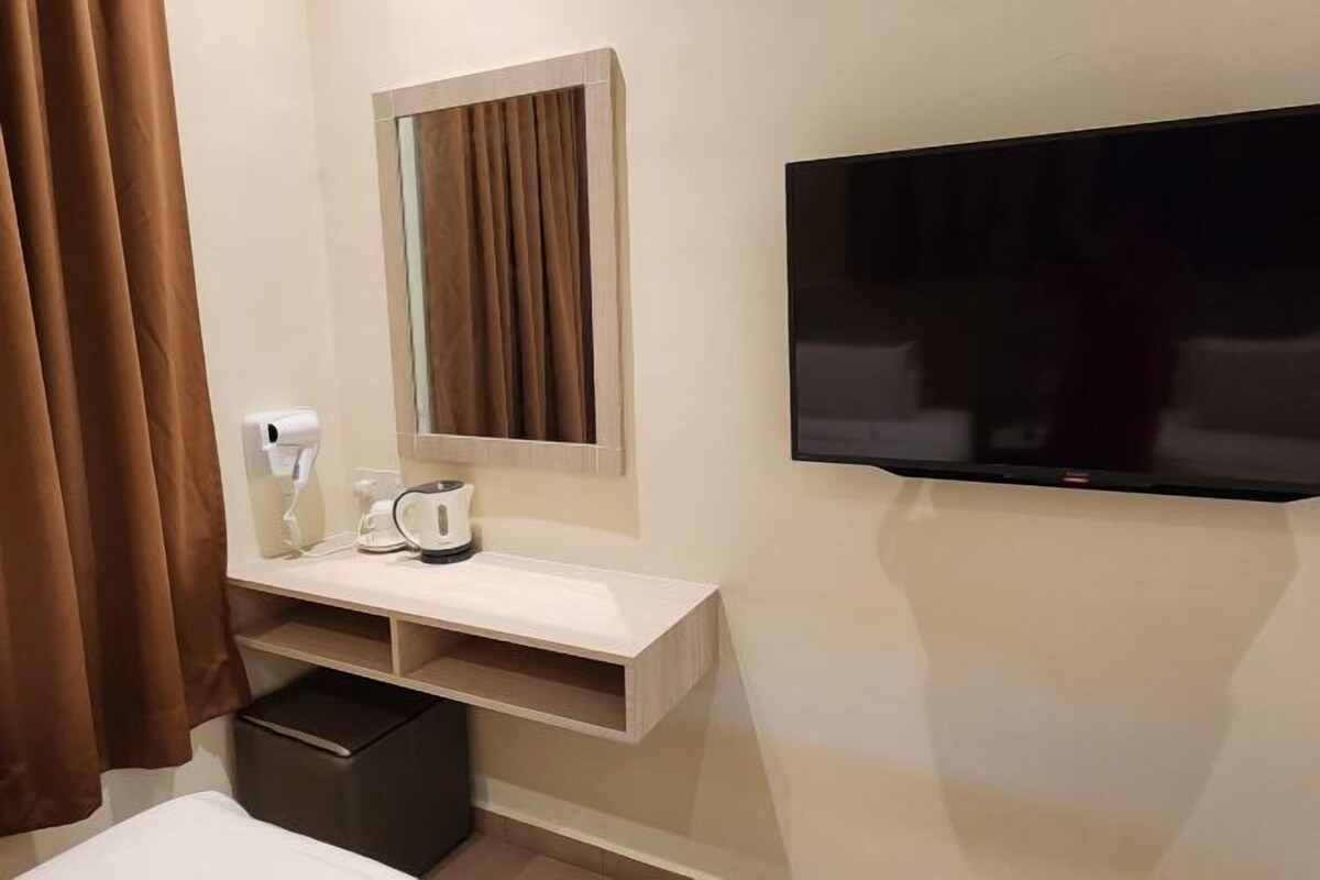 A hotel room with a TV mounted on the wall, a mirror above a vanity, a small shelf with toiletries, and a trash bin.