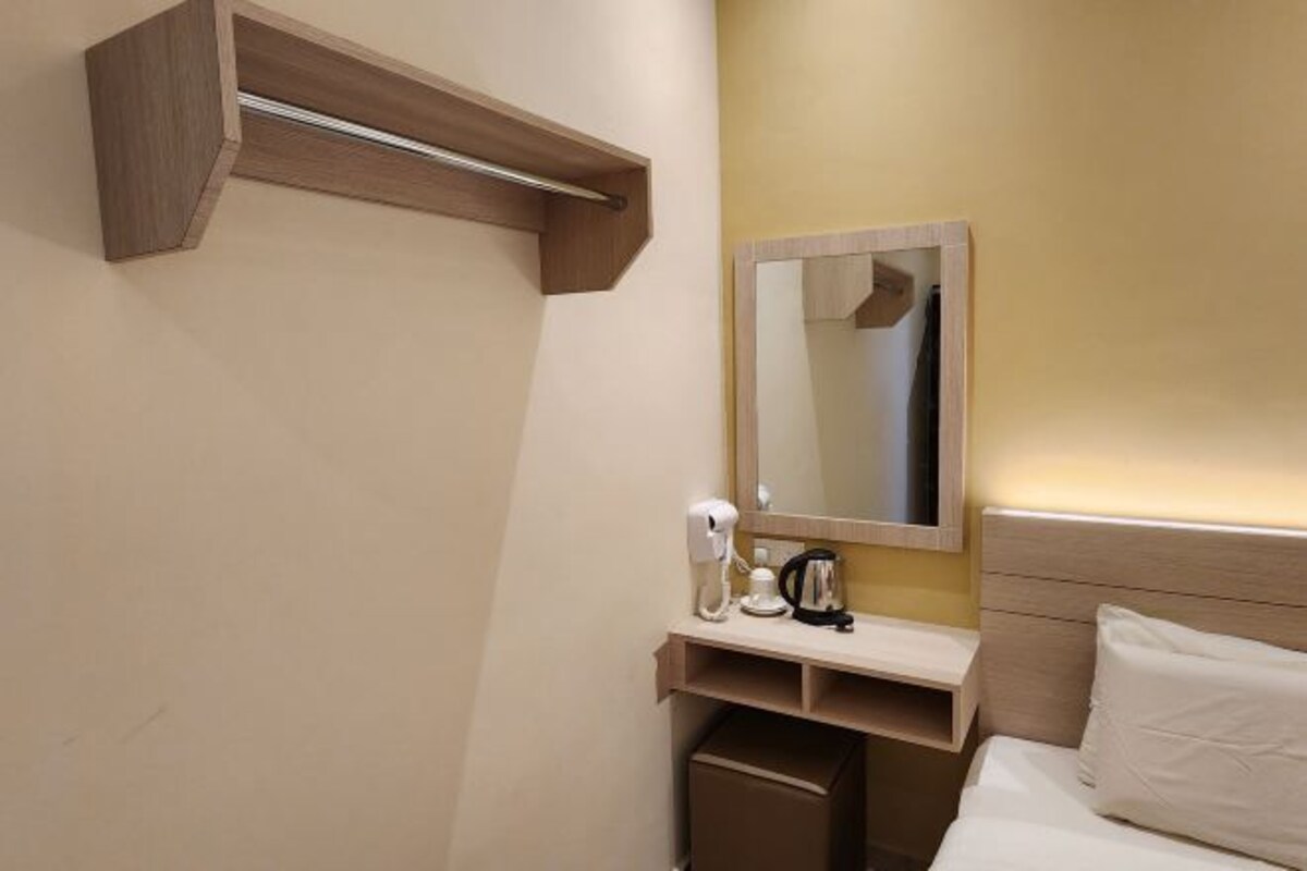A hotel room with a bed, a bedside table with a coffee maker, a mirror, and a wooden shelf.