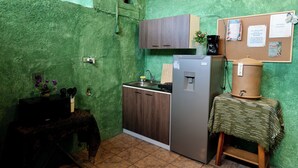 A compact kitchen with a green textured wall, a wooden cabinet, a stainless steel refrigerator, and a small table with a cloth. - Wonderland Hostel (Antigua Guatemala)