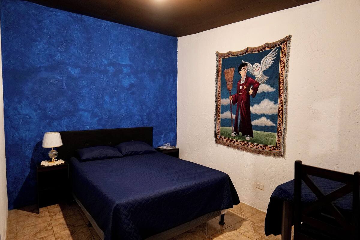 Ein Schlafzimmer mit blauer Wandstruktur, einem Bett mit dunkelblauer Bettdecke, einem Nachttisch mit Lampe und einem gerahmten Bild eines Zauberers an der Wand.