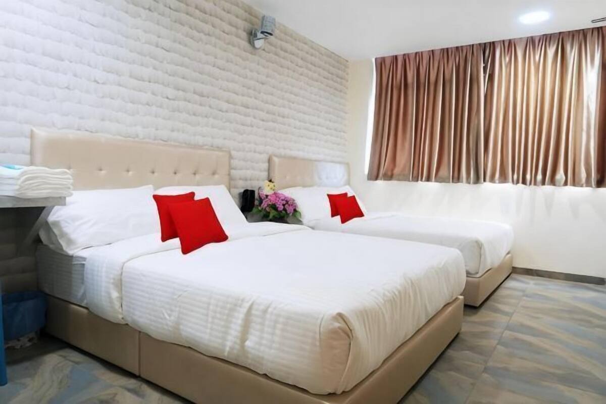 A hotel room with two beds, a white textured wall, a window with curtains, and a small table with a vase of flowers.