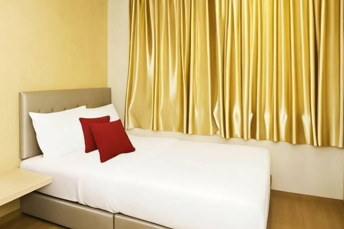 A bed with white bedding and a red pillow, a bedside table, and a curtain.