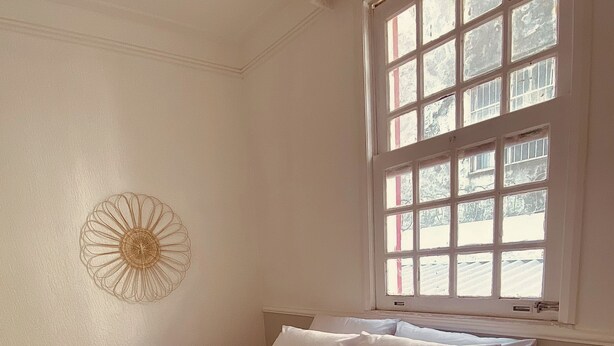 Ein ordentlich bezogenes Bett in einem Zimmer mit großem Fenster, einem kleinen Nachttisch und einer Wanddekoration.