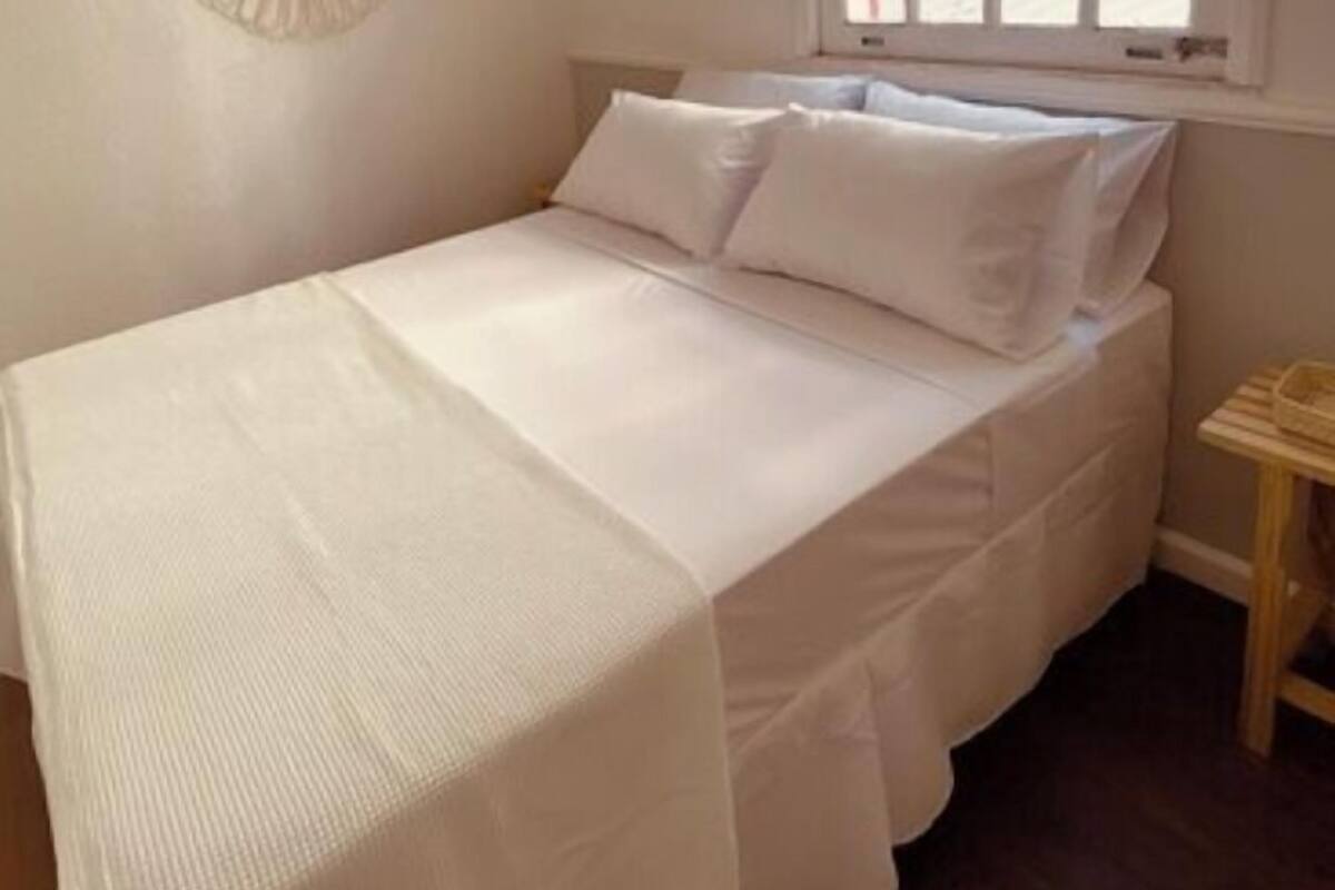 Ein Zimmer mit einem Einzelbett, einer schlichten weißen Bettdecke, einem Wanddekor und einem Fenster mit Blick auf ein Gebäude.
