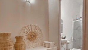 Ein Badezimmer mit weißem Waschbecken, WC und Dusche. Auf dem Regal befindet sich ein Korbwägelchen und ein dekorativer Fächer.