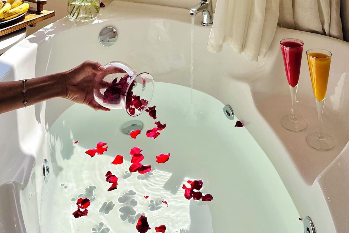 A bathtub with rose petals and two glasses of red and orange beverages, a vase of red roses, and a view of a pyramid through a window.