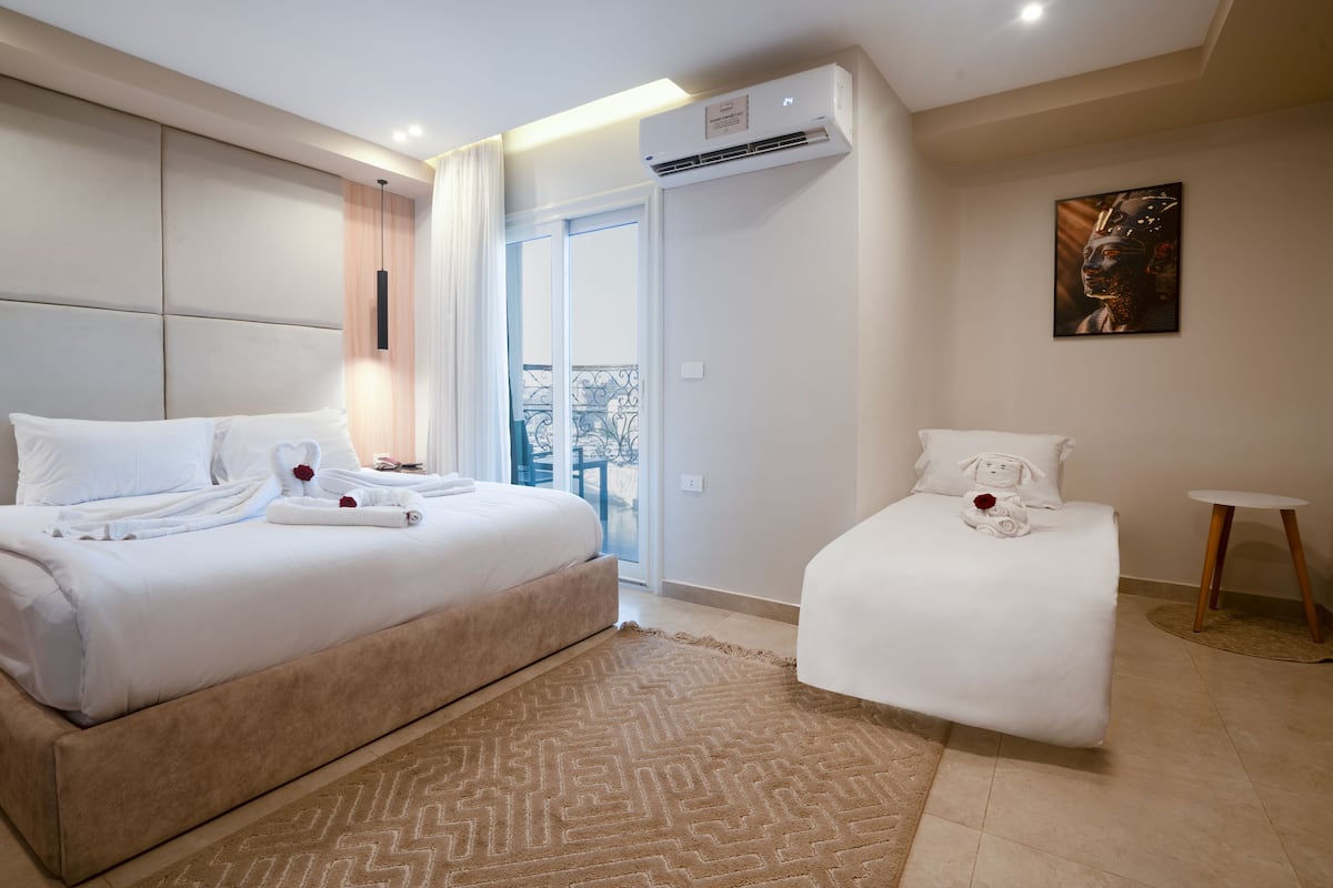 A hotel room with a large bed, a smaller bed, a balcony with a view, and an air conditioning unit.