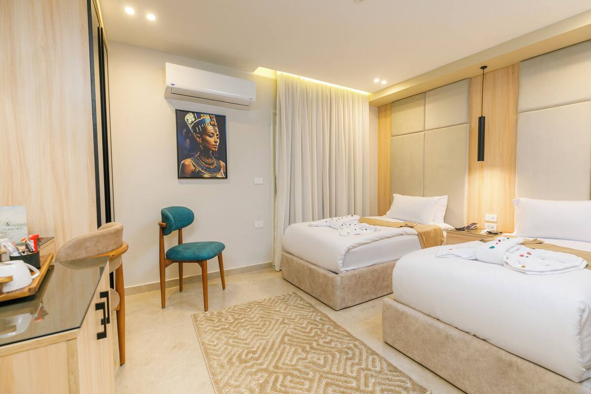 A hotel room with two beds, a desk, a chair, and a painting on the wall.