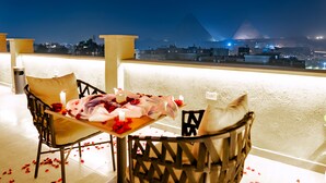 A balcony with a view of the pyramid at night, a table with rose petals, and two chairs. - Tiye Pyramids Hotel (Giza)