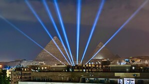 A pyramid with blue light beams converging towards it, surrounded by buildings with satellite dishes. - Tiye Pyramids Hotel (Giza)