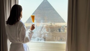 A person in a white bathrobe standing by a window with a view of a pyramid. - Tiye Pyramids Hotel (Giza)