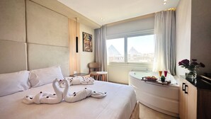 A hotel room with a bed, a bathtub, a view of the pyramid, and a vase with flowers. - Tiye Pyramids Hotel (Giza)